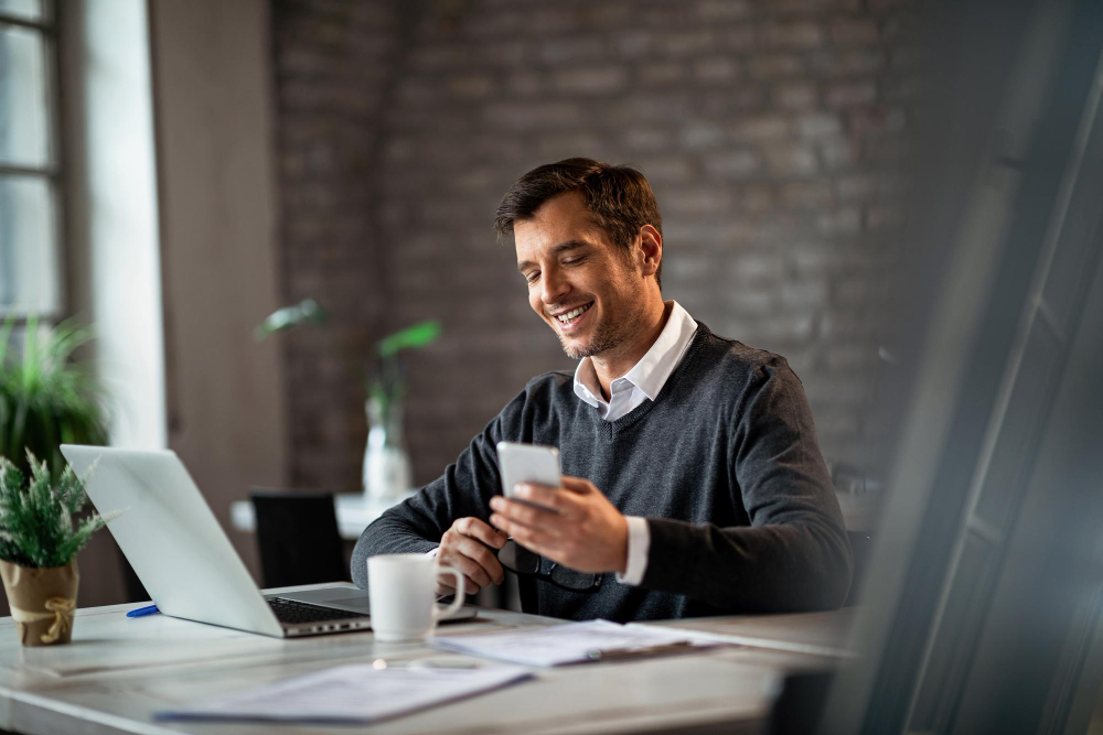 happy businessman texting on mobile phone while working on laptop in the office